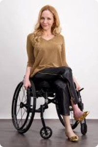 Woman in a wheelchair is cross legged wearing black trousers with side seam fastening and beige v neck jumper